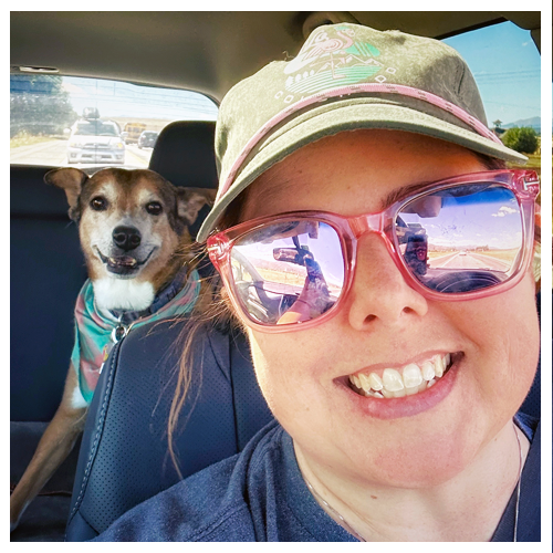 Woman in sunglasses and baseball cap in driver's seat of car with smiling rescue mutt in the backseat.