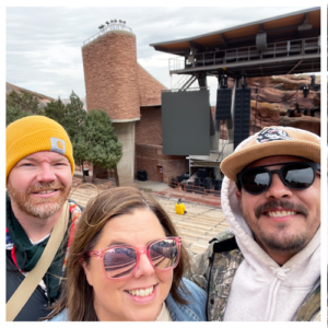 A trio of friends in sunglasses and winter gear in front of the stage at Red Rocks