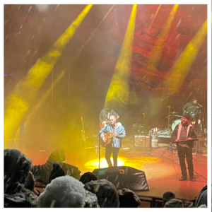 Zach Bryan on stage at Red Rocks under yellow and orange lights while heavy snow falls